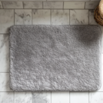 Dense grey chenille bath mat on white rental apartment tile, warm morning bathroom light from above, the pile texture visible and soft, the rubber backing edge slightly visible, moody editorial home photography, no people