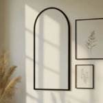 A large blank white rental apartment wall with a single powder-coated black arched mirror mounted to the left and botanical art prints in simple black frames, soft warm afternoon light from a window, moody editorial apartment photography, no people