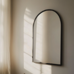 A powder-coated matte black arched mirror mounted on a plain white rental apartment wall, warm afternoon light reflected in the tempered glass, the rest of the wall still bare, soft shadow beneath the arch frame, moody editorial apartment photography, no people