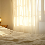 Soft spring morning light coming through a gap in white sheer curtains in a minimal rental apartment bedroom, a neatly made bed partially visible, warm ambient light from a nightstand lamp, the room dim and quiet, editorial bedroom photography, cream and ivory and pale morning light palette, no people