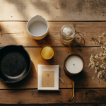 Flat lay of five apartment essentials on warm wood surface: cast iron skillet, ceramic V60 dripper, tobacco patchouli candle, matte gold desk lamp, and lint roller, editorial interior photography, natural light, warm amber tones